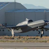 Silver and black experimental aircraft landing near industrial hangar with mountains in background