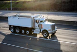 White Peterbilt dump truck traveling on a multi-lane highway with grassy hillside in background