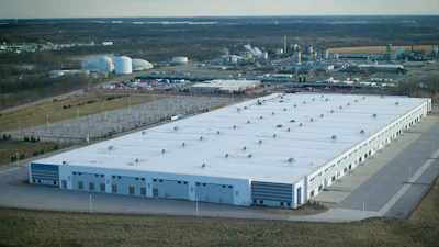 Aerial view of large industrial warehouse facility with white roof and blue walls in rural landscape