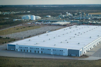 Aerial view of large industrial warehouse facility with white roof and blue walls in rural landscape